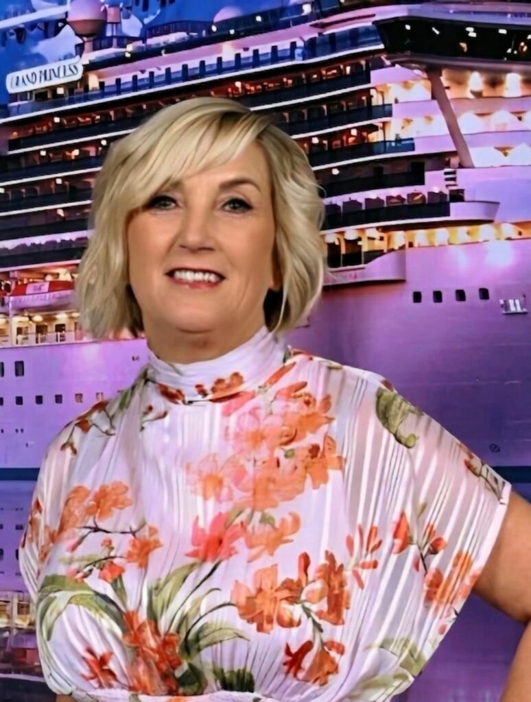 A woman with short blonde hair, Master Stylist and Owner, poses in a floral dress before a large cruise ship illuminated with purple and pink lighting.