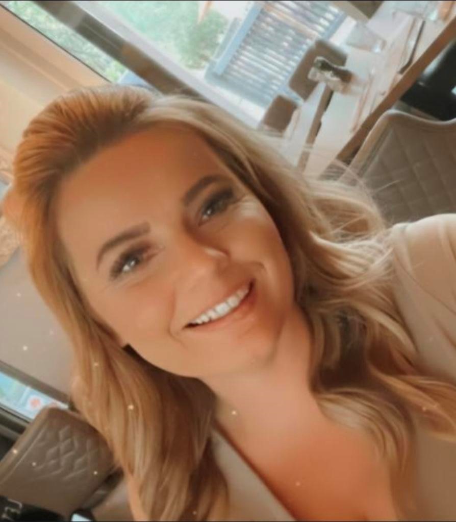Rachel, a salon manager with long blonde hair, smiles at the camera as she sits indoors at a table surrounded by quilted chairs, with windows visible in the background.