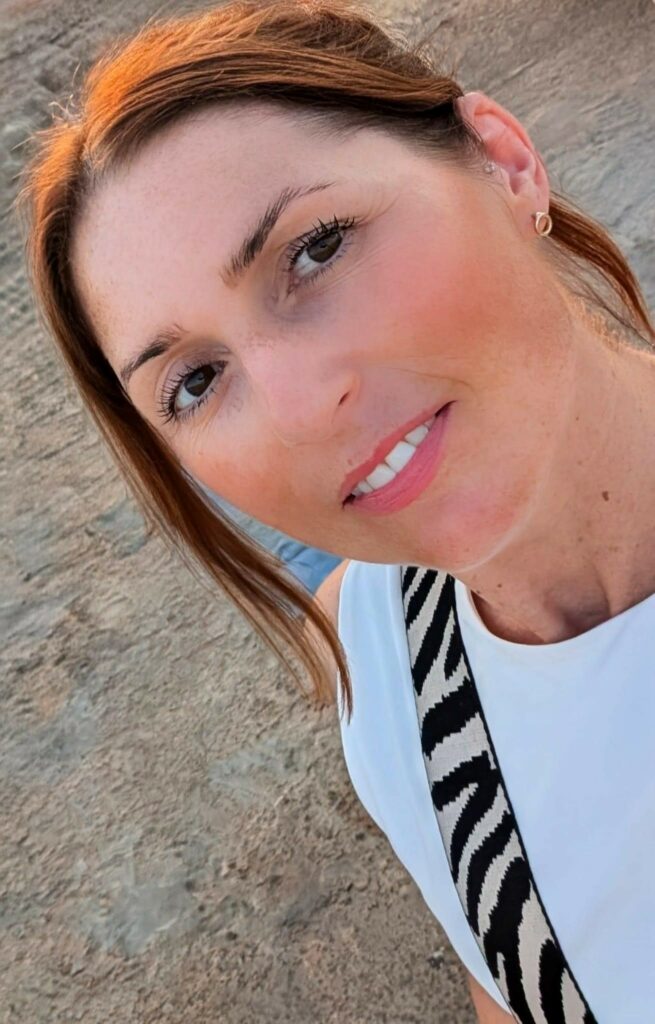 Kirsty Graham, a beauty therapist with fair skin and brown hair, takes a close-up selfie outdoors. She wears a white top and a black-and-white striped bag strap, with sand visible in the background.