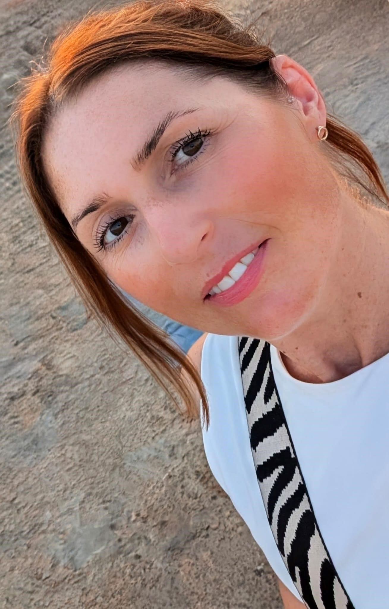 Kirsty Graham, a beauty therapist with fair skin and brown hair, takes a close-up selfie outdoors. She wears a white top and a black-and-white striped bag strap, with sand visible in the background.
