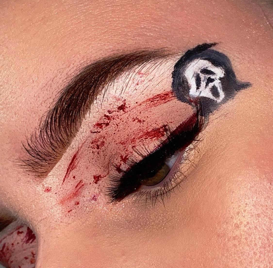 Close-up of an eye adorned with striking makeup, showing smeared red pigment, a touch of macadamia shimmer, and a tiny black-and-white ghost face motif by the outer corner of the eyelid.