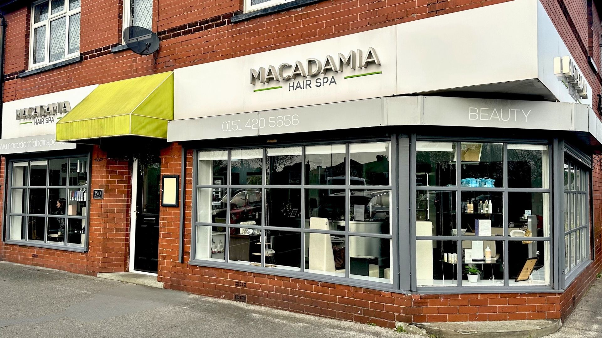 Street view of Macadamia Hair Spa, a brick corner building with large glass windows displaying Macadamia hair and beauty products, and a yellow canopy above the entrance.