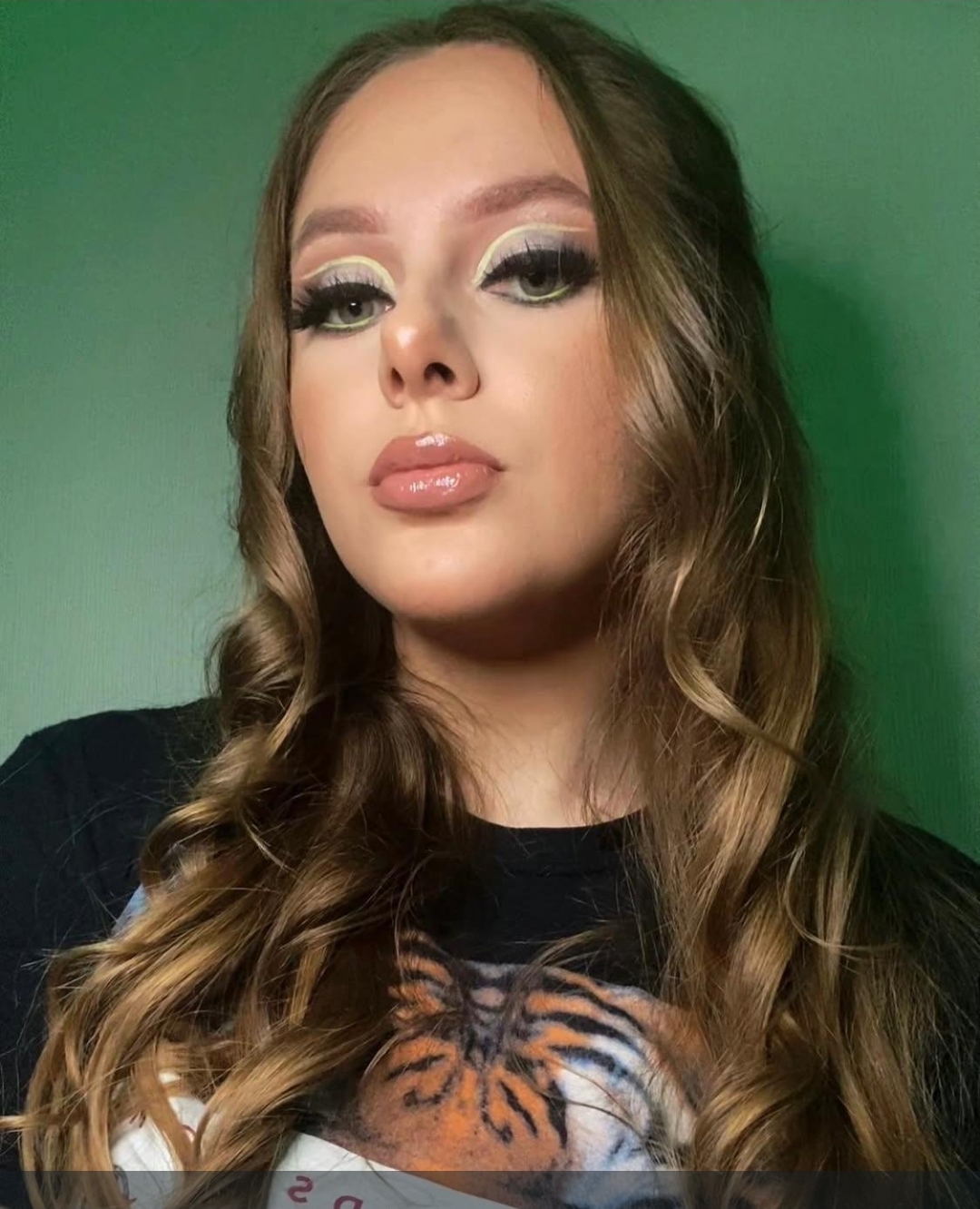 A young woman with long, wavy hair—styled like a proper Salon Support Supervisor—poses against a green background. She’s wearing make-up and a black top featuring a tiger design.