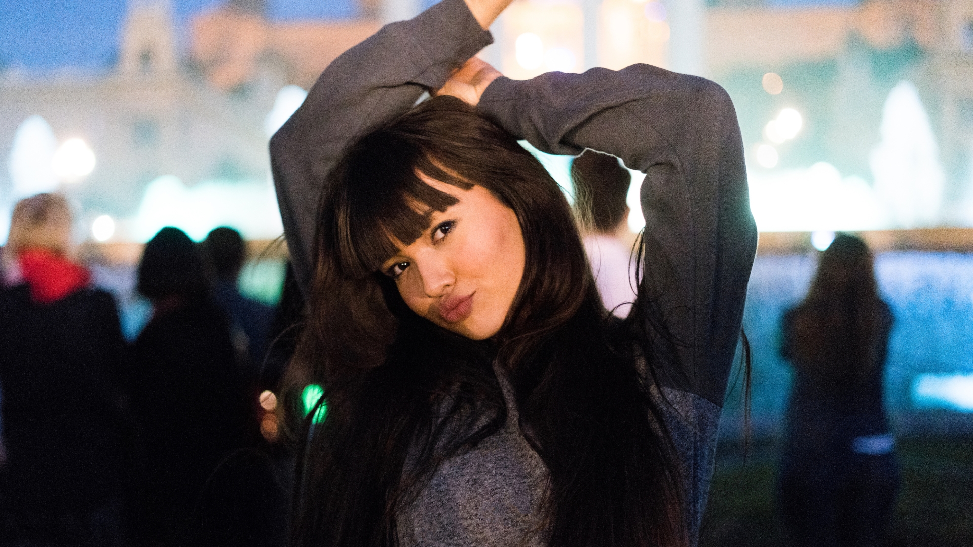 A woman with long, dark hair and fashionable fringe poses outdoors at night with her arms raised and lips pursed, showcasing the latest Spring 2025 hair trends amidst blurred figures and bright lights.