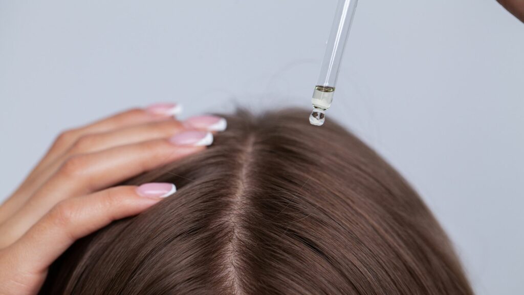 A hand parts brown hair while another holds a dropper, applying liquid to the scalp to promote healthy hair this spring.