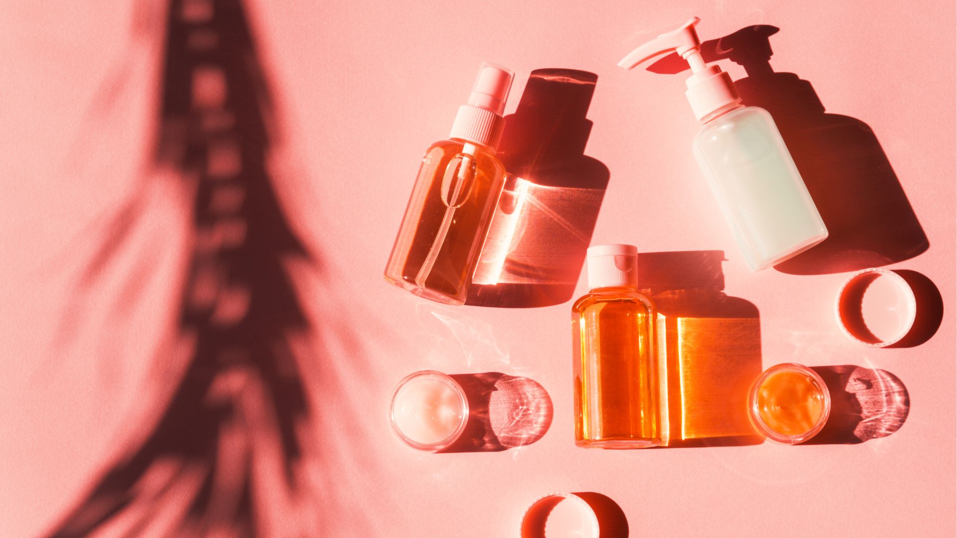 A selection of skincare bottles and jars containing creams and liquids, arranged on a pink surface alongside hair preparation essentials, with pronounced shadows and the silhouette of a palm leaf.
