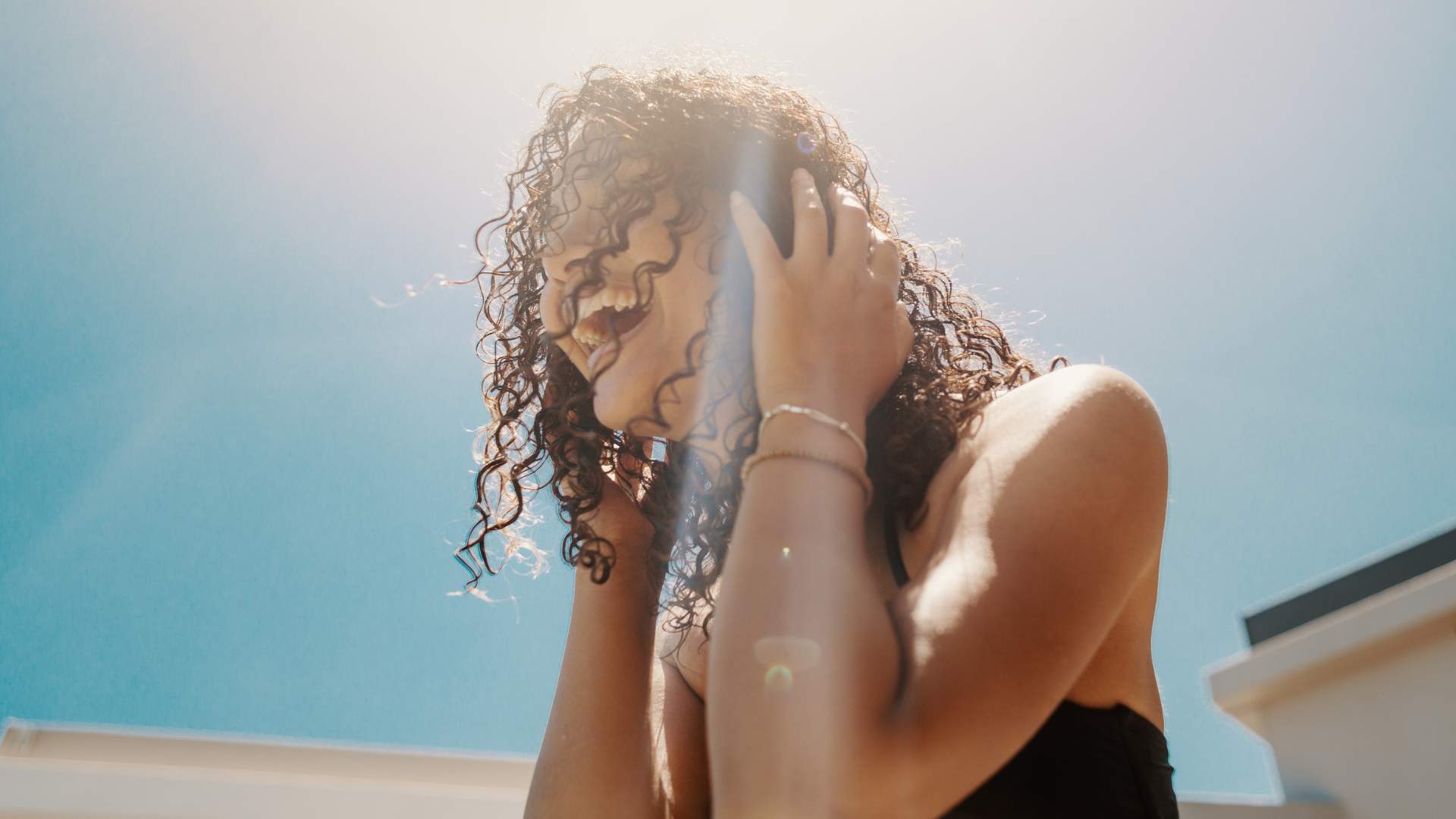 A person with curly hair smiles while holding their head, enjoying their summer hair care routine outdoors beneath bright sunlight and a clear blue sky.