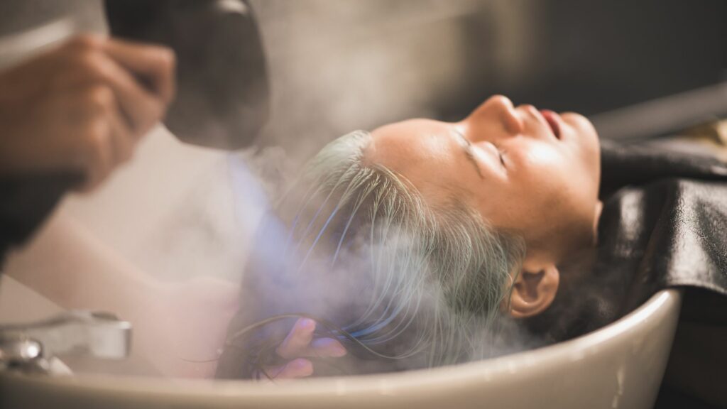 A person is enjoying a Micro Mist Hair Treatment at a salon, reclining with their eyes closed at the basin while steam is gently applied for deep nourishment of the hair.