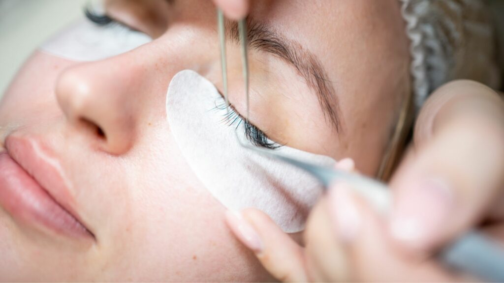 Close-up of a person having eyelash extensions applied, with tweezers placing the lashes and a protective pad beneath the eye—ideal for complementing healthy hair and a fresh summer hairstyle.