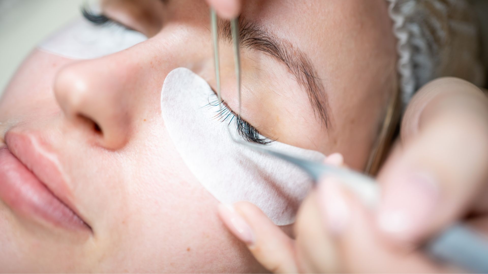 Close-up of a person having eyelash extensions applied, with tweezers placing the lashes and a protective pad beneath the eye—ideal for complementing healthy hair and a fresh summer hairstyle.