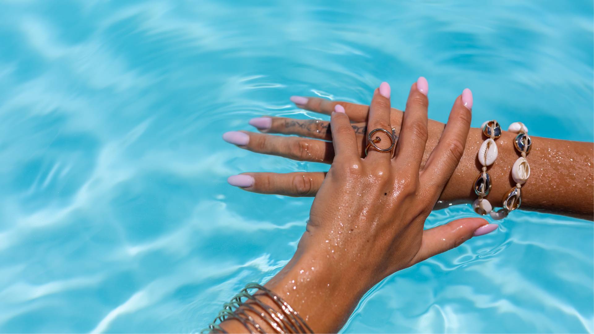 A close-up of hands with pink manicured nails, adorned with rings and shell bracelets, resting by a blue swimming pool—highlighting the latest summer nail trends.