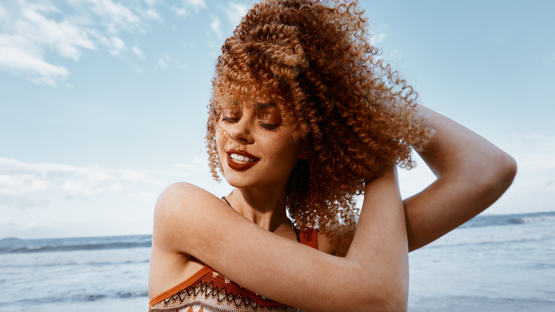 A woman with curly hair smiles with her eyes closed by the sea, her arms raised and hands running through her hair, embracing summer 2025 and the latest hair colour trends. The sky is partly cloudy.