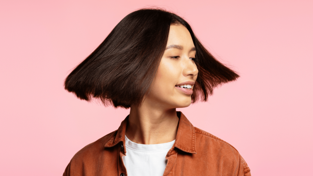 A person with straight, dark chin-length hair turns their head, causing their hair to fan out—a fresh look inspired by summer hairstyles. They are wearing a rust-coloured jacket over a white shirt, set against a pink background.
