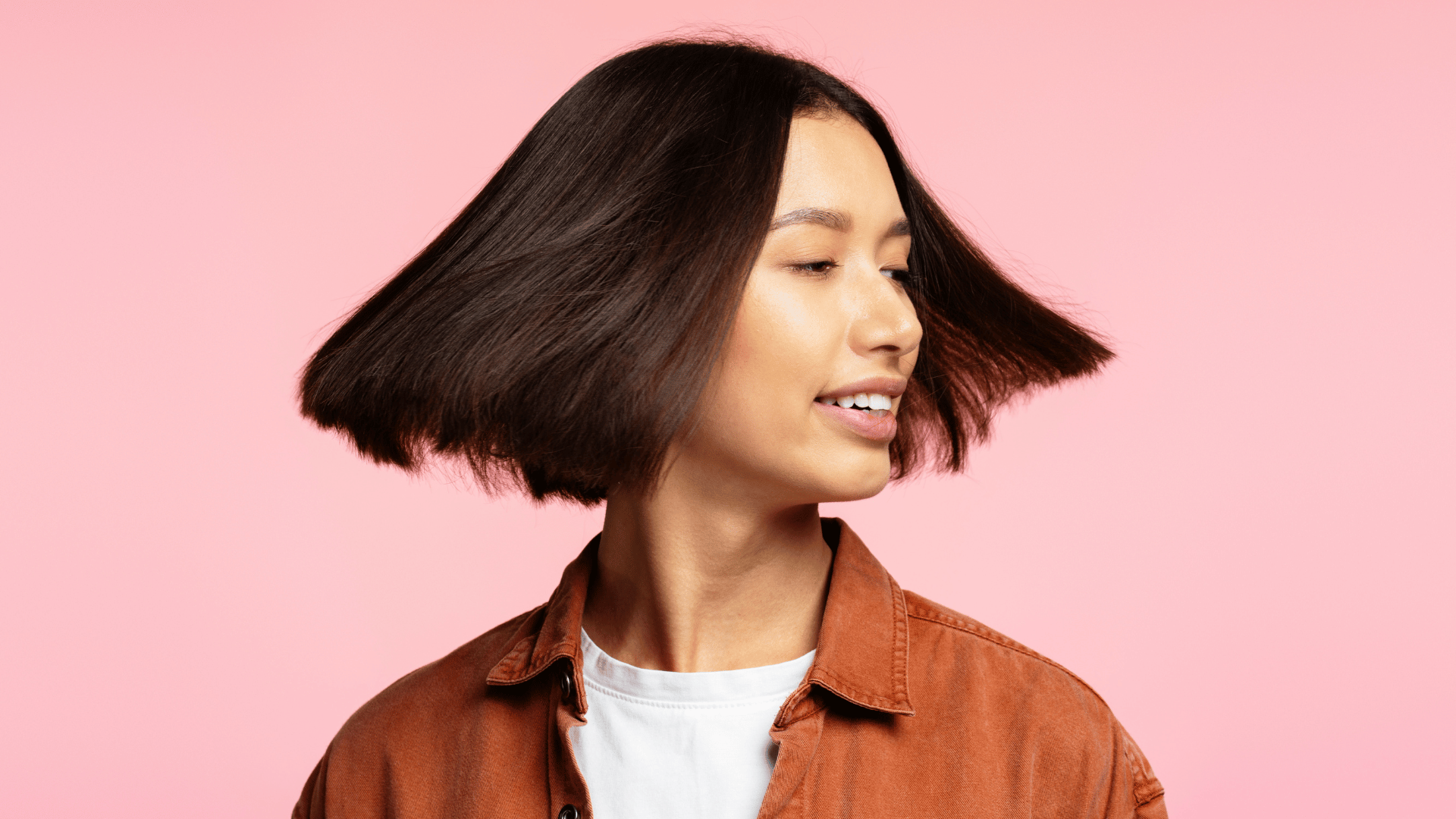 A person with straight, dark chin-length hair turns their head, causing their hair to fan out—a fresh look inspired by summer hairstyles. They are wearing a rust-coloured jacket over a white shirt, set against a pink background.
