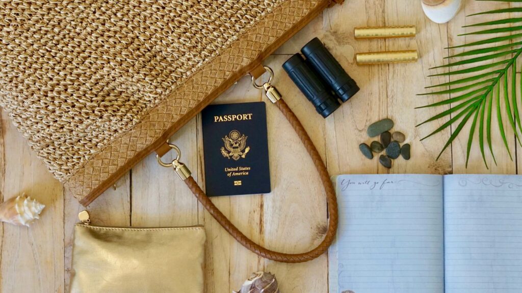 Woven handbag with a US passport, binoculars, notebook, gold pouch, seashells, pebbles and a palm leaf on a wooden surface—perfect for your next holiday adventure.