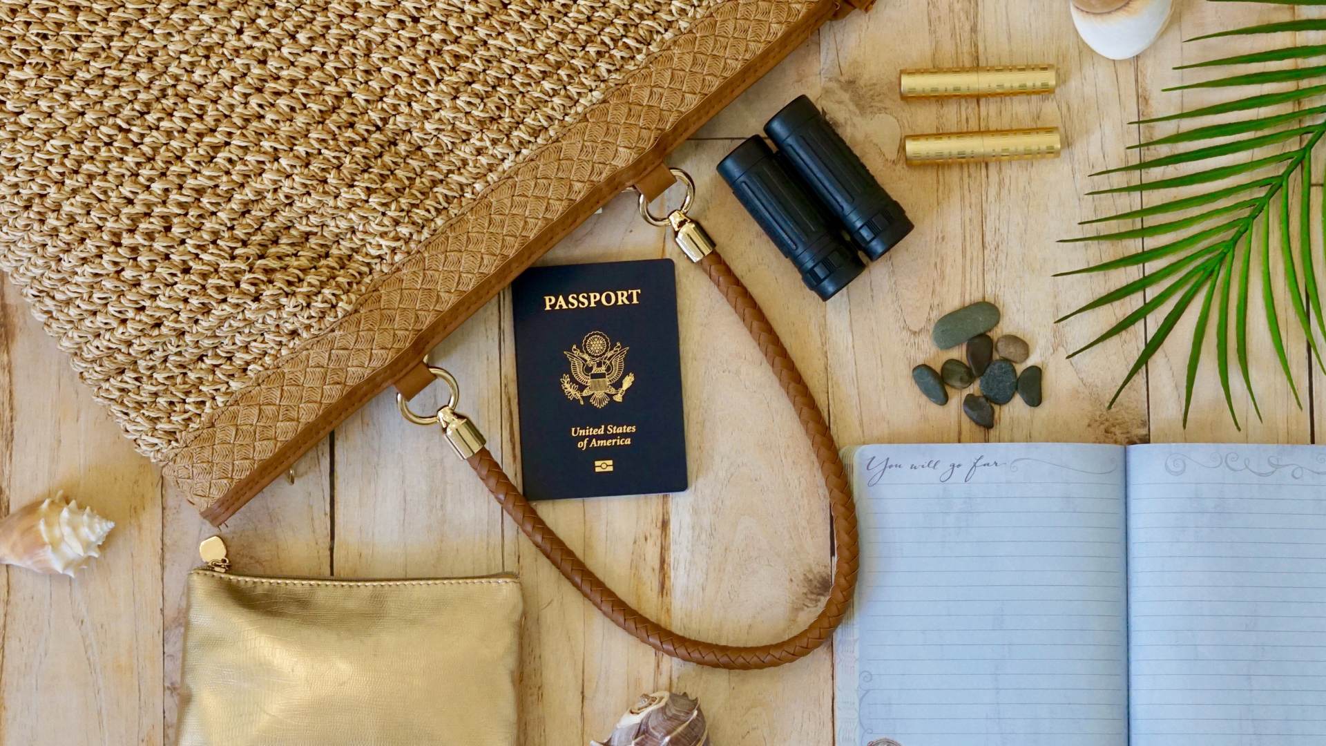 Woven handbag with a US passport, binoculars, notebook, gold pouch, seashells, pebbles and a palm leaf on a wooden surface—perfect for your next holiday adventure.