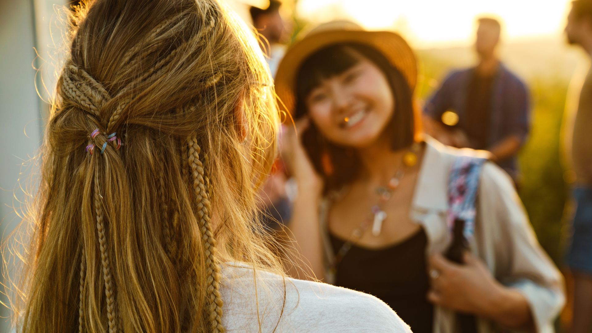 Two people are outdoors at sunset, with one woman’s festival hair in focus from behind and another smiling in the background, holding a bottle. Several others are blurred in the distance, exuding festival hairstyle vibes.