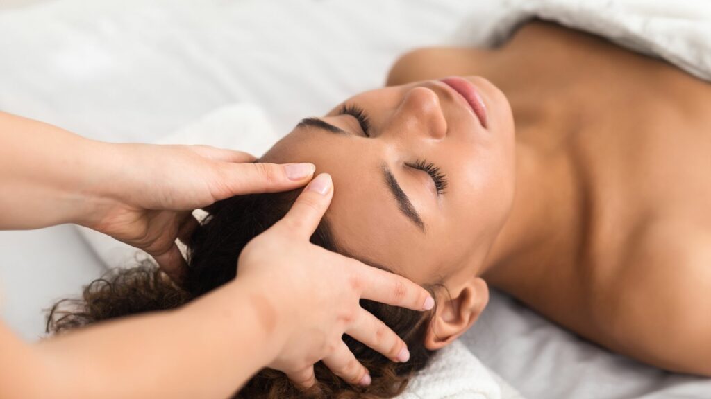 A person is lying down with their eyes closed, receiving a head massage for stress relief from another individual, covered with a white towel.