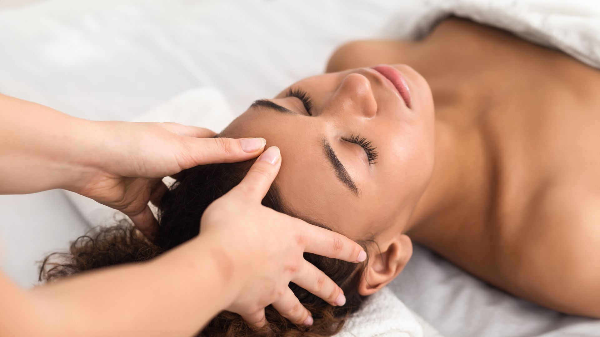 A person is lying down with their eyes closed, receiving a head massage for stress relief from another individual, covered with a white towel.