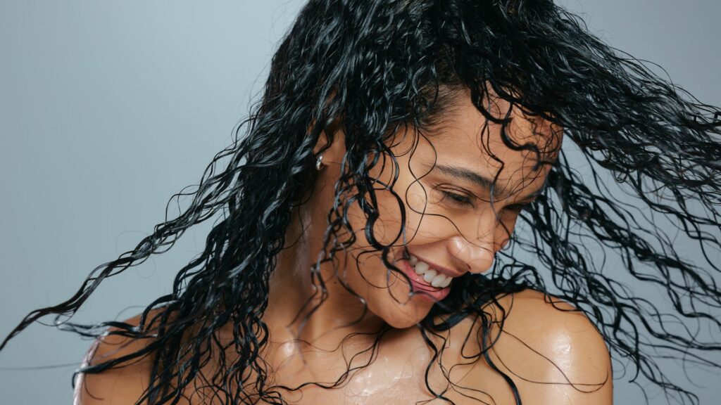 A woman with wet, curly hair smiles as she turns her head, with water droplets visible on her skin—demonstrating healthy hair care—set against a plain light grey background.