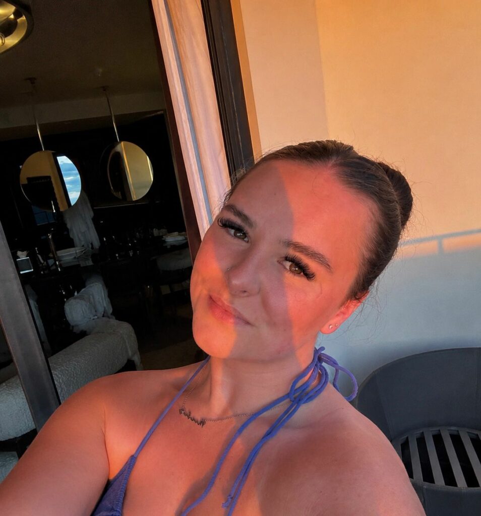 A woman, styled reminiscent of Phoebe by a skilled stylist, takes a selfie on a balcony at sunset. Sunlight and shadows play across her face as she wears a blue halter top and her hair is tied up in a bun.