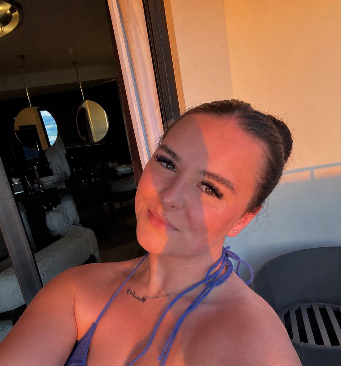 A woman, styled reminiscent of Phoebe by a skilled stylist, takes a selfie on a balcony at sunset. Sunlight and shadows play across her face as she wears a blue halter top and her hair is tied up in a bun.