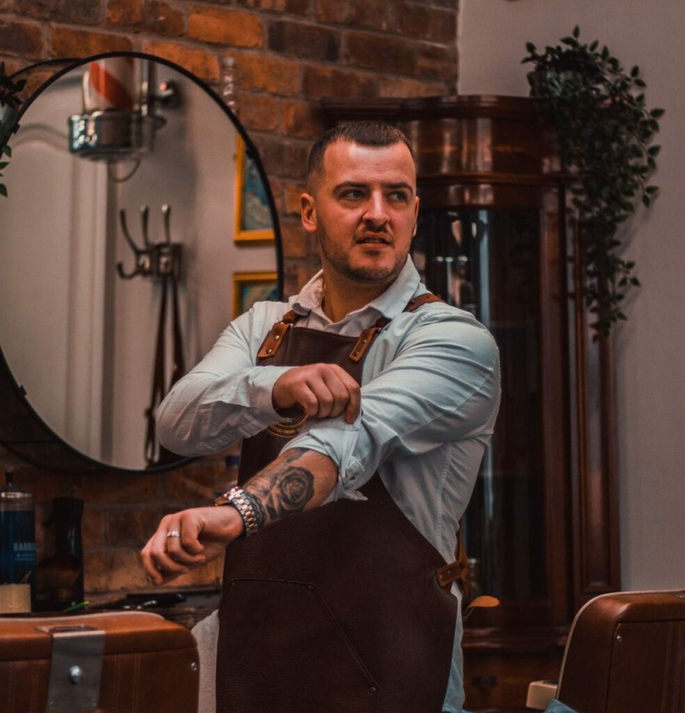 A barber wearing an apron rolls up his sleeve as he stands in a barbershop featuring brick walls, mirrors, and barber chairs.