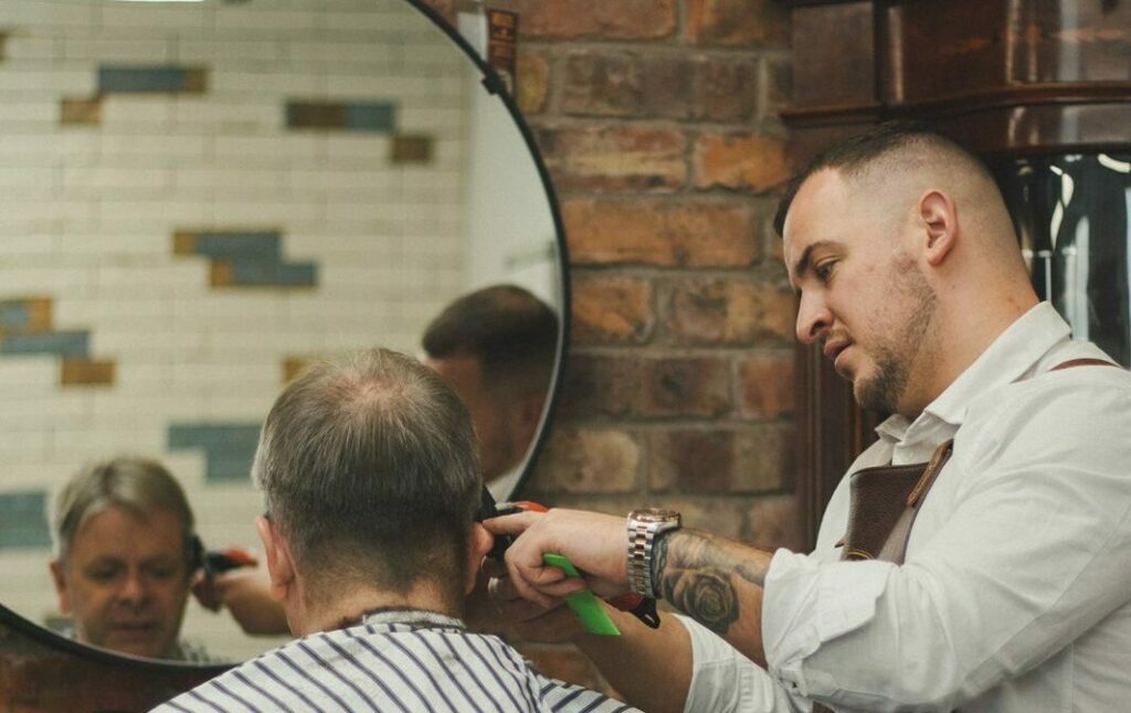 A barber wearing an apron cuts the hair of a client seated with a striped cape in a barbershop featuring a brick wall and a round mirror.
