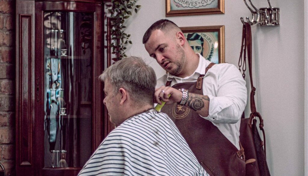 A barber in an apron trims a seated customer’s hair in a barbershop adorned with framed portraits and a traditional striped barber’s pole.