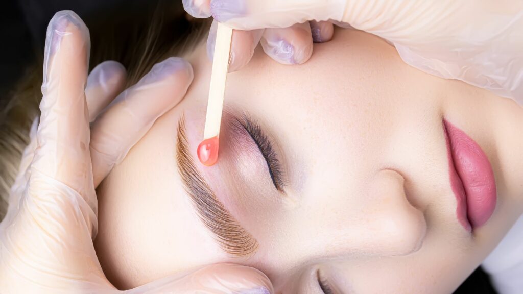 Close-up of a person with eyes closed having their eyebrows waxed; gloved hands use a wooden spatula to apply wax above the eyebrow for gentle hair removal and a smooth finish.