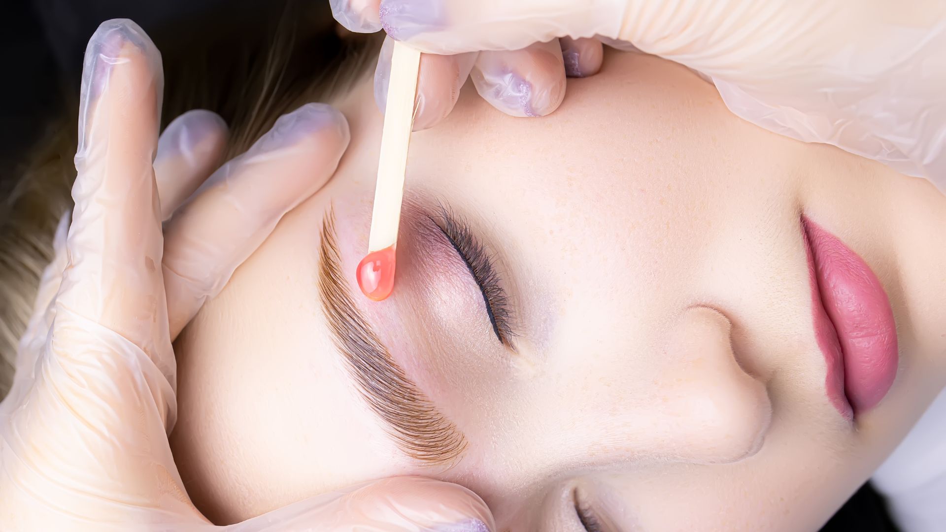 Close-up of a person with eyes closed having their eyebrows waxed; gloved hands use a wooden spatula to apply wax above the eyebrow for gentle hair removal and a smooth finish.