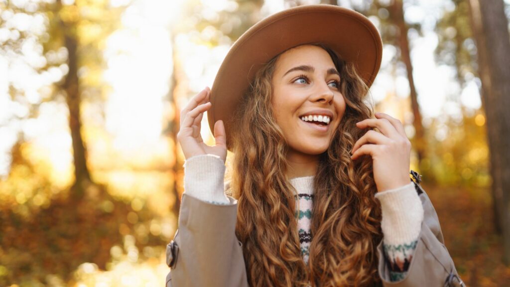 A woman with long curly hair, wearing a hat and a patterned jumper, smiles while standing outdoors in a sunlit woodland, highlighting autumn fashion amidst the vibrant colours of the season.