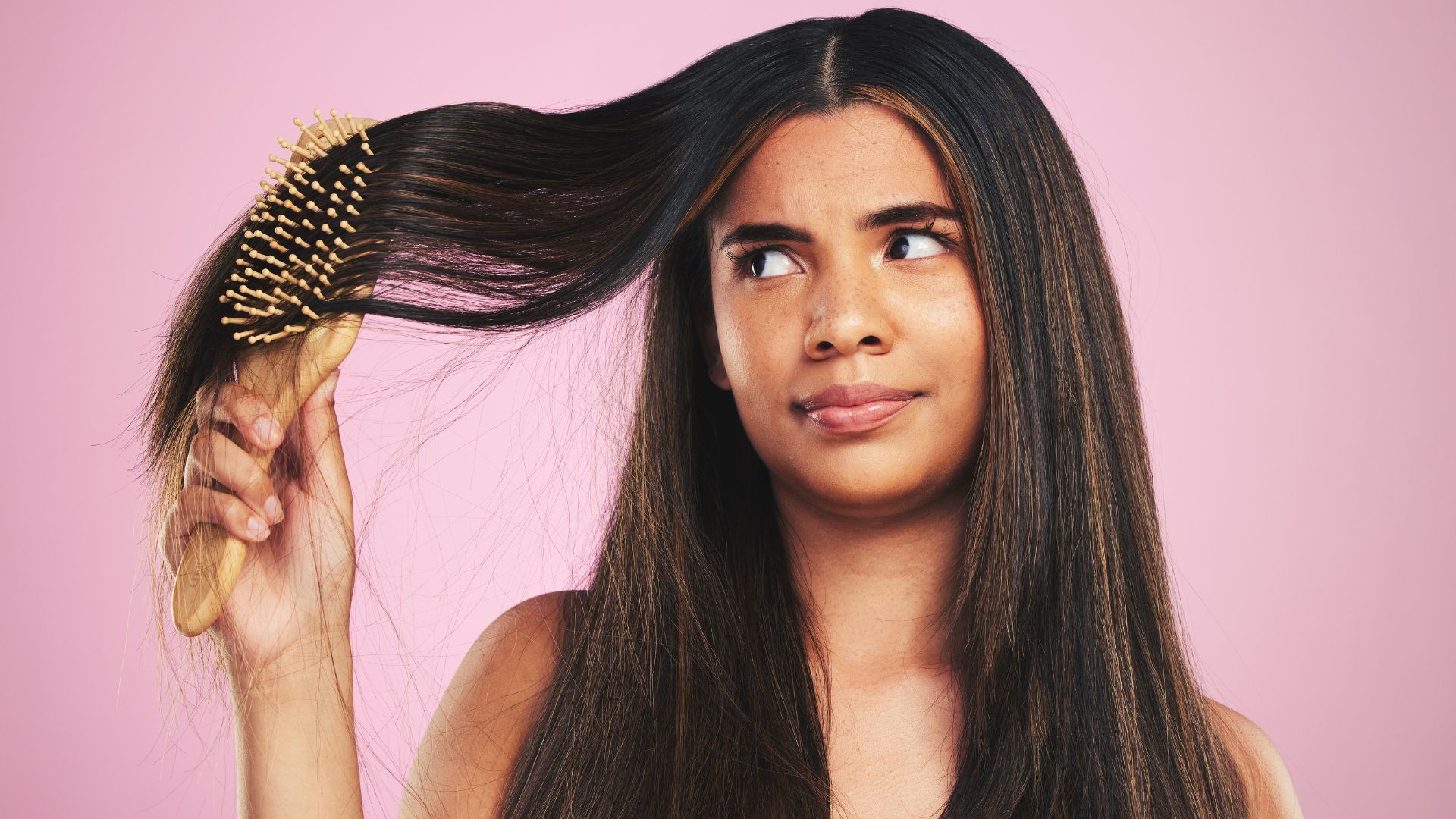 A woman with long dark hair holds a hairbrush caught in her hair, gazing at it with a worried expression against a pink background—its truly a hair SOS situation.