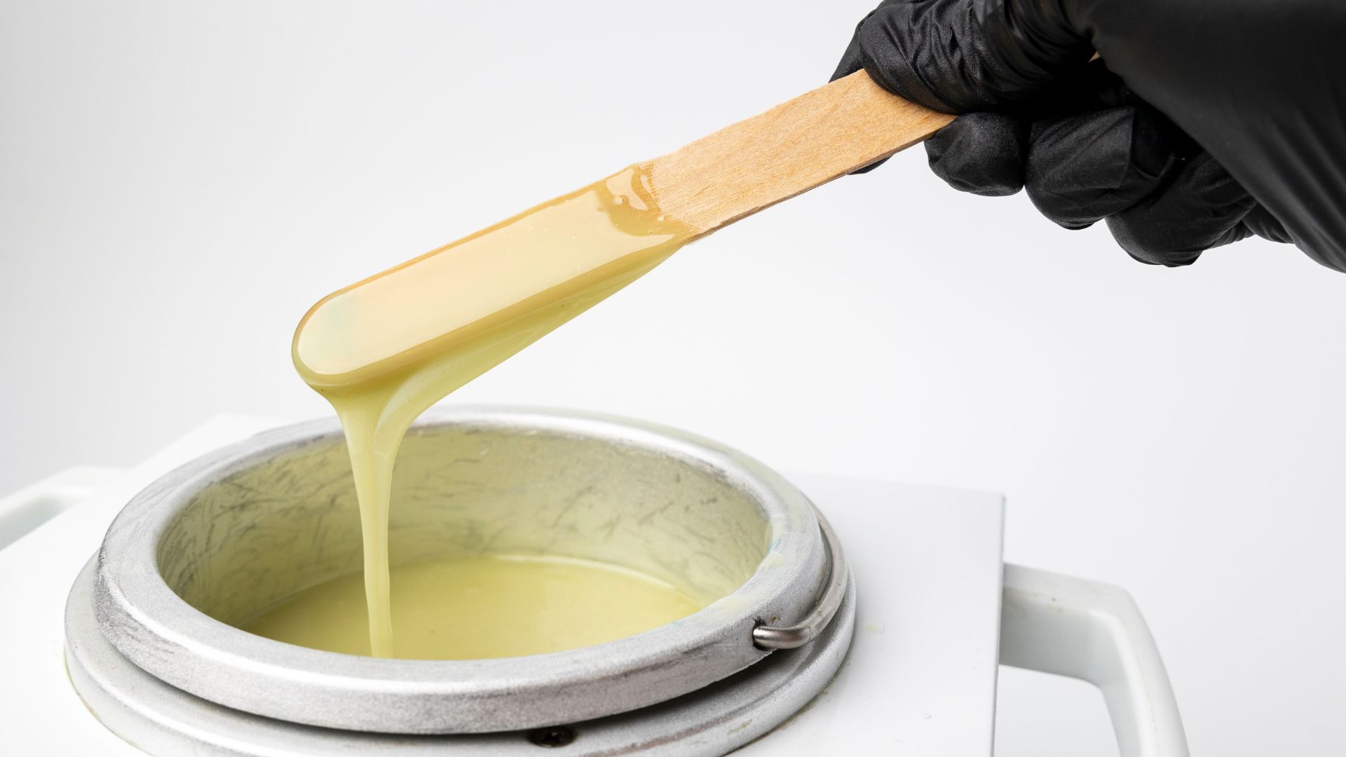 A gloved hand holds a wooden spatula with warm wax dripping from it above a metal wax warmer, prepared for smooth skin and efficient hair removal.