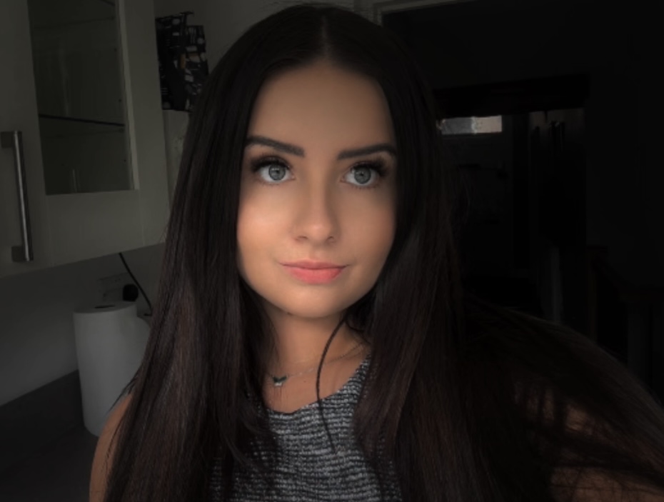 A young woman with long dark hair and blue eyes, a Senior Stylist, gazes slightly away from the camera while standing indoors in a softly lit room.