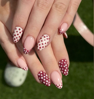 A hand with almond-shaped autumn nails, painted with a pink base and decorated with red and white polka dots, shown against a green background—a fun take on seasonal nail art.