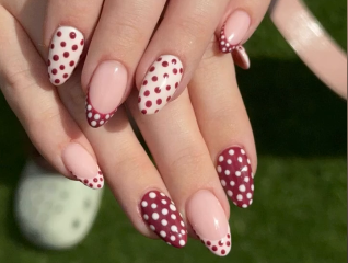 A hand with almond-shaped autumn nails, painted with a pink base and decorated with red and white polka dots, shown against a green background—a fun take on seasonal nail art.