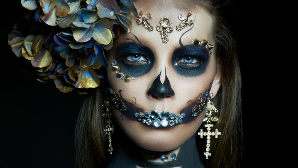Individual adorned with intricate Day of the Dead face paint, immaculate makeup, sparkling jeweled decorations, a large floral arrangement in their hair, and ornate cross earrings, gazing directly at the camera.
