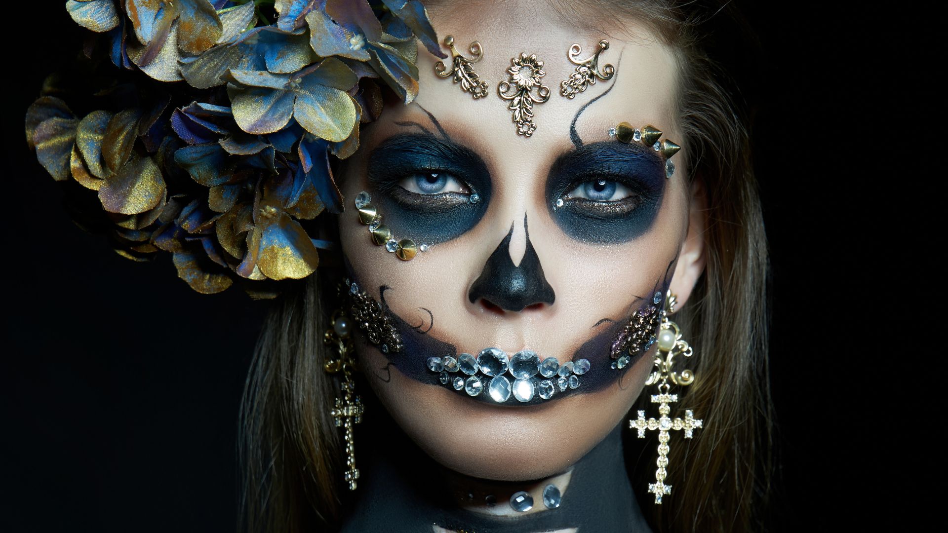 Individual adorned with intricate Day of the Dead face paint, immaculate makeup, sparkling jeweled decorations, a large floral arrangement in their hair, and ornate cross earrings, gazing directly at the camera.