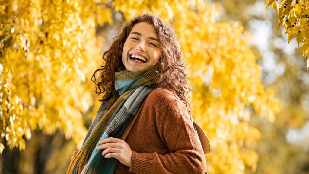 A person with curly hair and a scarf enjoys the cosy season, smiling outside in front of trees adorned with yellow autumn leaves.