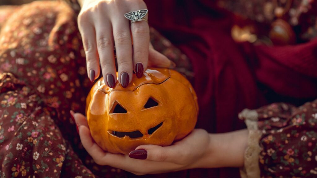 Someone wearing a floral dress holds a small carved pumpkin with a jack-o'-lantern face; their hand, adorned with maroon Halloween nails and a butterfly ring, rests on the top.