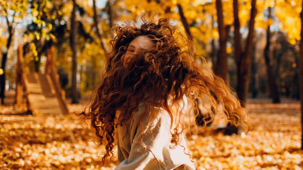 Individual with long, curly hair twirling outside in a sun-dappled woodland, autumn leaves carpeting the ground, enjoying seasonal hair care for lively, revitalised tresses.
