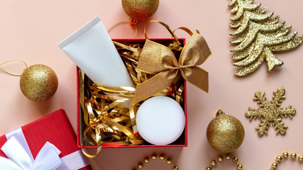 A red Christmas gift box with a gold ribbon holds a white tube and jar, surrounded by gold baubles, a gold tree, and a snowflake—ideal for gifting beautiful hair this season, set against a peach background.