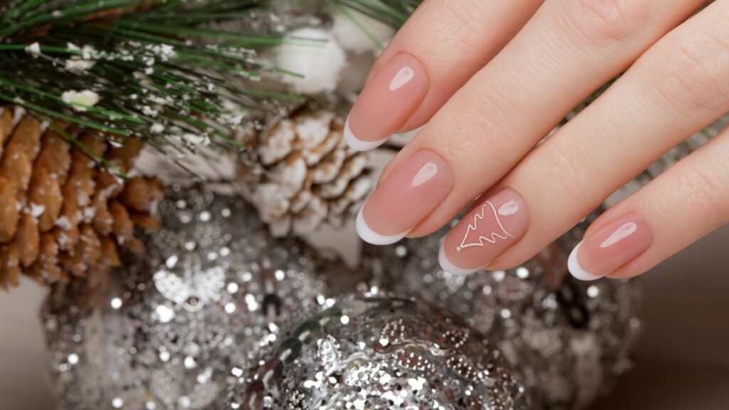 A hand with French manicured nails—one featuring a festive white outline of a Christmas tree—rests beside pine cones, pine needles, and sparkling silver baubles.