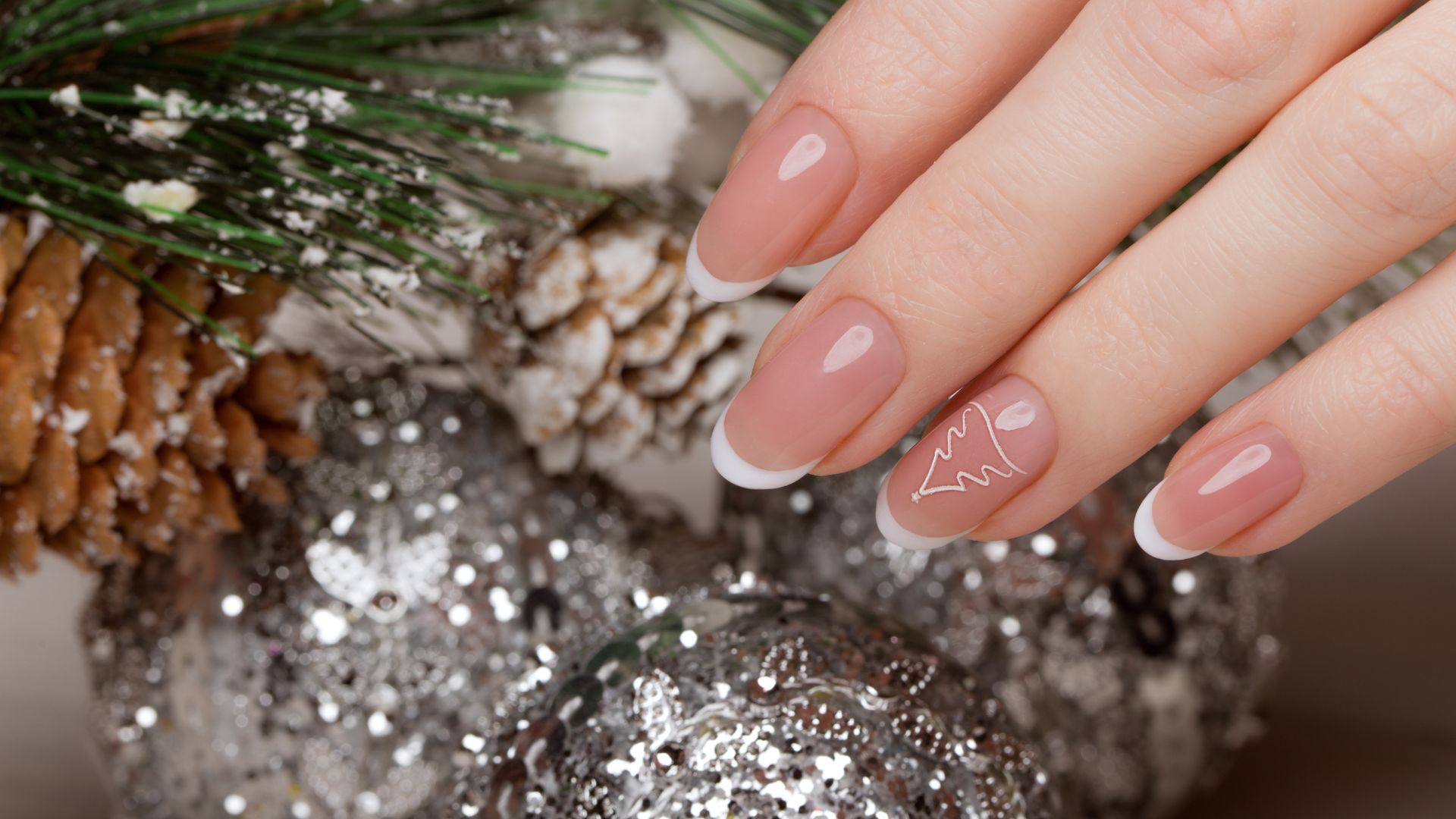 A hand with French manicured nails—one featuring a festive white outline of a Christmas tree—rests beside pine cones, pine needles, and sparkling silver baubles.