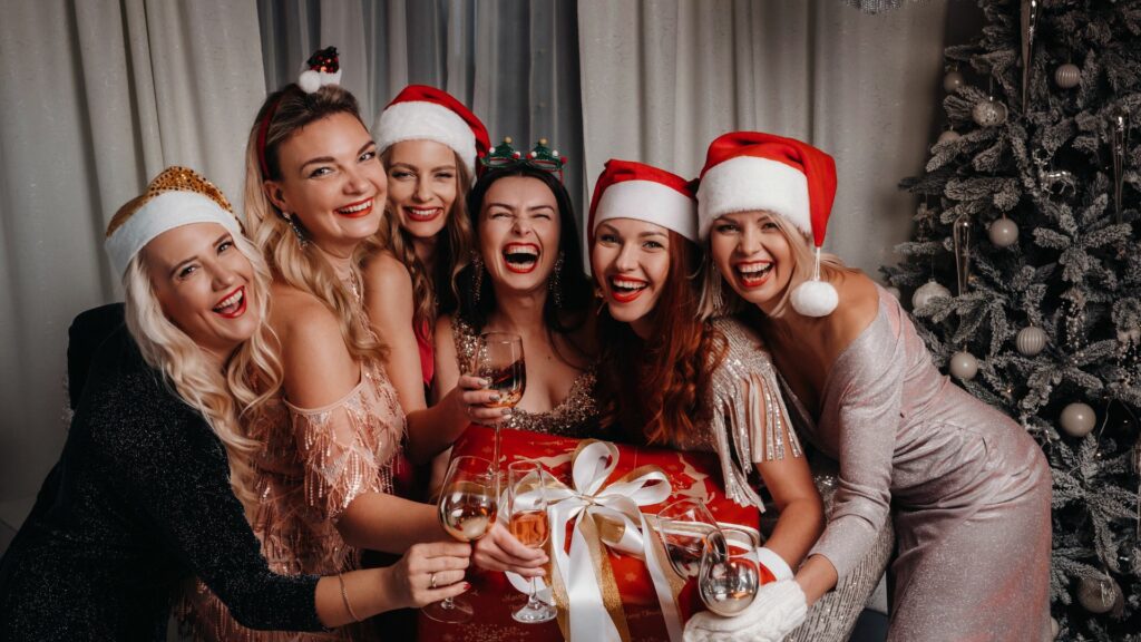 Six women with beautiful hair, dressed in festive outfits and Santa hats, smile as they hold glasses and a Christmas gift box in front of a decorated tree.
