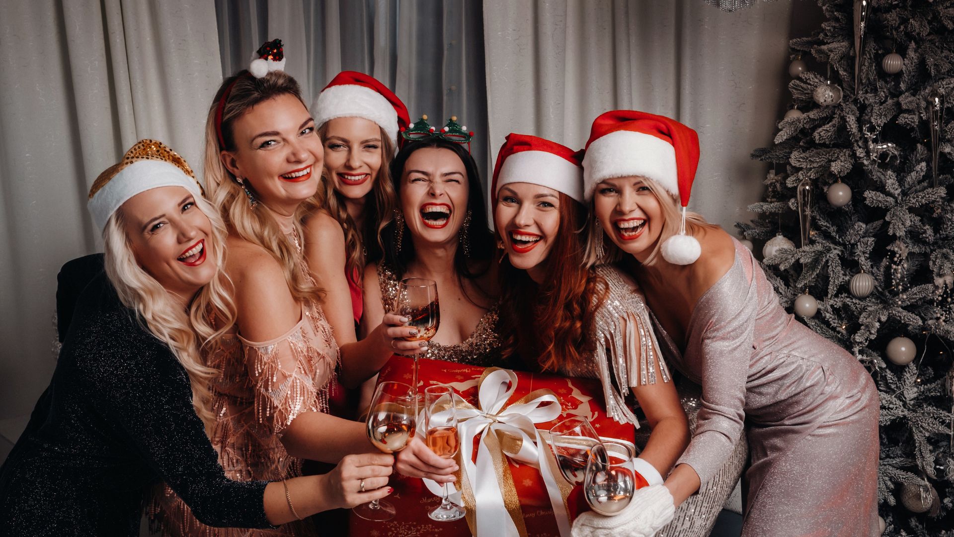 Six women with beautiful hair, dressed in festive outfits and Santa hats, smile as they hold glasses and a Christmas gift box in front of a decorated tree.