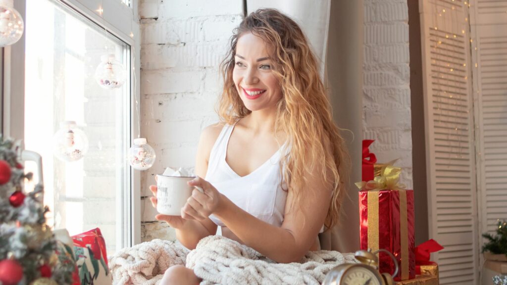 A woman sits by a window, smiling as she holds a mug, surrounded by wrapped presents and festive Christmas decorations—enjoying a moment of Christmas self-care as the holiday celebrations draw nearer.