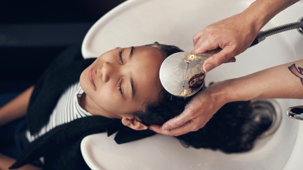 A child reclines with eyes closed at a salon basin as an adult washes their hair using a handheld sprayer, enjoying a relaxing hair care experience.