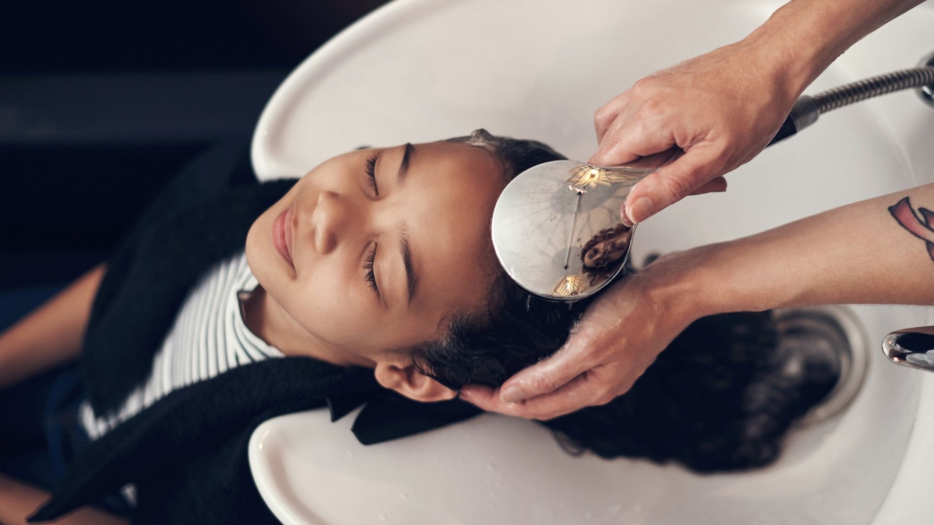 A child reclines with eyes closed at a salon basin as an adult washes their hair using a handheld sprayer, enjoying a relaxing hair care experience.